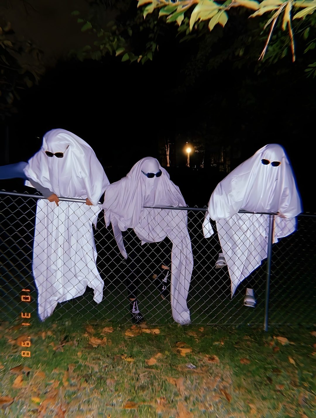 three white ghost costumes hanging on a fence in the dark, with one person standing next to them Trio Halloween black