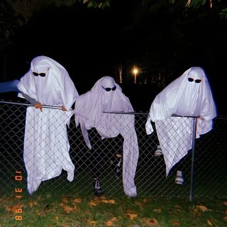 three white ghost costumes hanging on a fence in the dark, with one person standing next to them Trio Halloween black