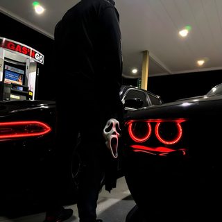 a man in a hoodie standing next to two cars with red lights on them Halloween Hellcat Halloween Car Decoration Ideas, Nighttime Gas Station Scene, Person With Mask Between Cars, black