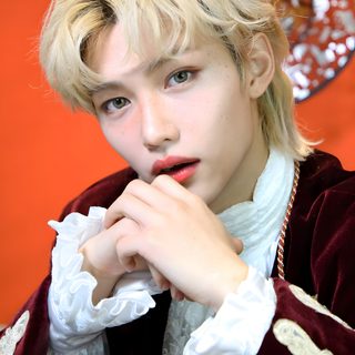 a young man with blonde hair wearing a red and white jacket sitting in front of an orange wall Felix Halloween Felix Costume, Felix Skz Halloween Costume, Felix Halloween Costume, indian red