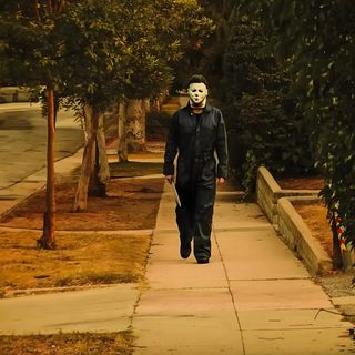 a man walking down a sidewalk with a mask on his face and the words john carpenter's John Carpenter Halloween John Carpenter, dark olive green