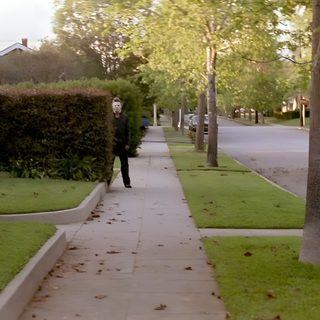 a man walking down a sidewalk next to a lush green park filled with trees and bushes Halloween 1978 Haddonfield Aesthetic, Midwest Halloween, Midwest Halloween Aesthetic, dark olive green