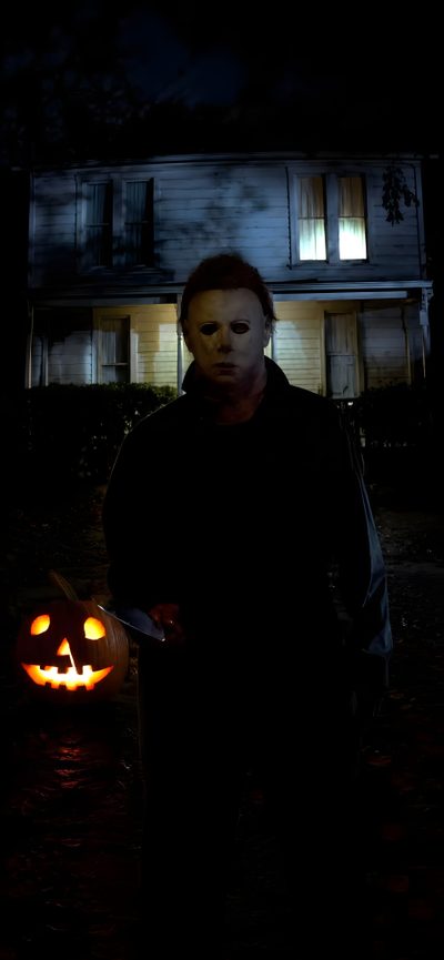a man standing in front of a house with two pumpkins on the ground and one person holding a knife Halloween The Movie Horror Iphone, Michael Myers, Horror Villains, Halloween, black