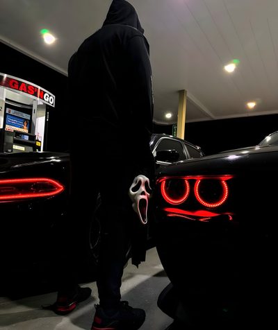 a man in a hoodie standing next to two cars with red lights on them Halloween Hellcat Halloween Car Decoration Ideas, Nighttime Gas Station Scene, Person With Mask Between Cars, black