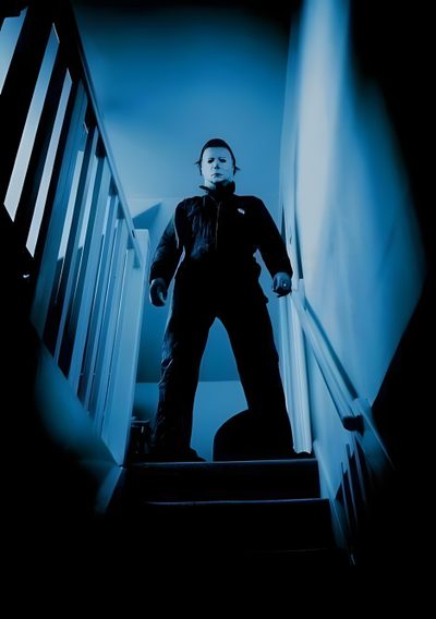 a man standing at the top of stairs in front of a stair case with blue light coming from behind him Halloween 1978 Michael Myers Halloween Aesthetic Michael Myers, Michael Myers In The Dark, Michael Myers 78, black