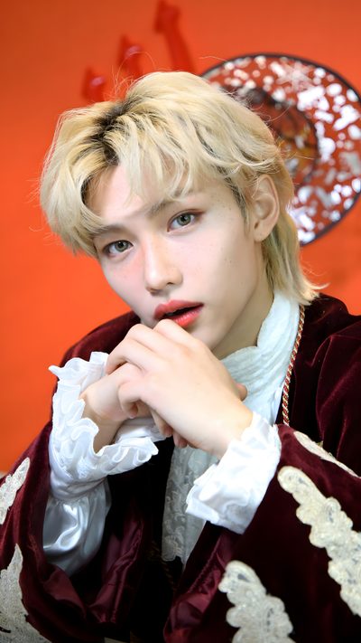 a young man with blonde hair wearing a red and white jacket sitting in front of an orange wall Felix Halloween Felix Costume, Felix Skz Halloween Costume, Felix Halloween Costume, indian red