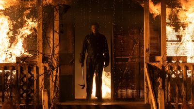 a man standing in front of a door with fire coming out of the building behind him Halloween 2018 Michael Myers Michael Myers Scenes, Michael Myers House On Fire, Michael Myers In Fire, maroon
