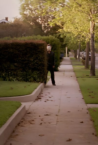 a man walking down a sidewalk next to a lush green park Halloween 1978 Horror Movie Scenes, The Babadook, Halloween 1978, Halloween Film, dark olive green
