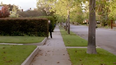 a man walking down a sidewalk next to a lush green park filled with trees and bushes Halloween 1978 Haddonfield Aesthetic, Midwest Halloween, Midwest Halloween Aesthetic, dark olive green
